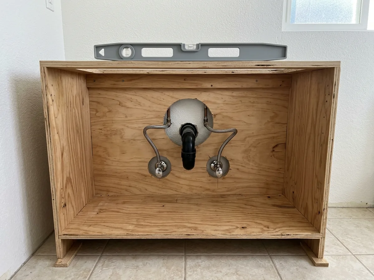 Plywood vanity base with clean cutouts in the back panel around a drain and two supply valves